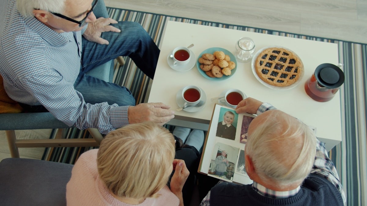 Group of elderly friends men and woman are watching pictures in photoalbum chatting sitting at table with food relaxing enjoying communication.